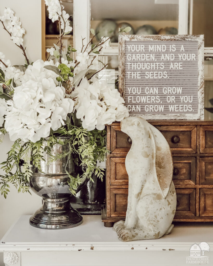 This springtime vignette includes a bunny statue, beautiful blooms, and a spring-themed letterboard quote